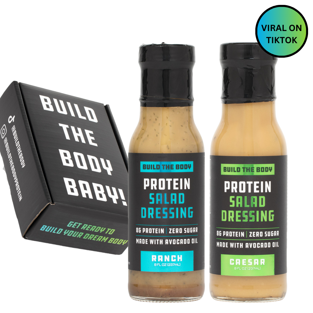Two bottles of 'Build the Body' protein salad dressing, flavors Caesar and Ranch, in a black box.
